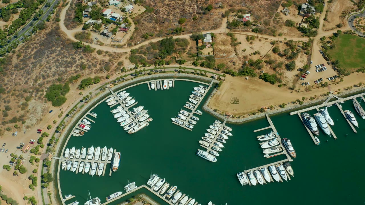 A 0:02 overhead frame of Puerto Los Cabos marina shows compact boat rows on green water, a curved shoreline wall, dry scrubland, roadways, and no visible people.