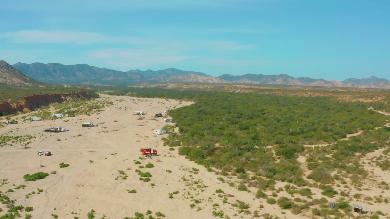 Opening aerial frame over Los Frailes Los Cabos showing a red truck on pale sand, scattered vehicles, green shrubs, distant mountains, clear blue sky, and no visible people.