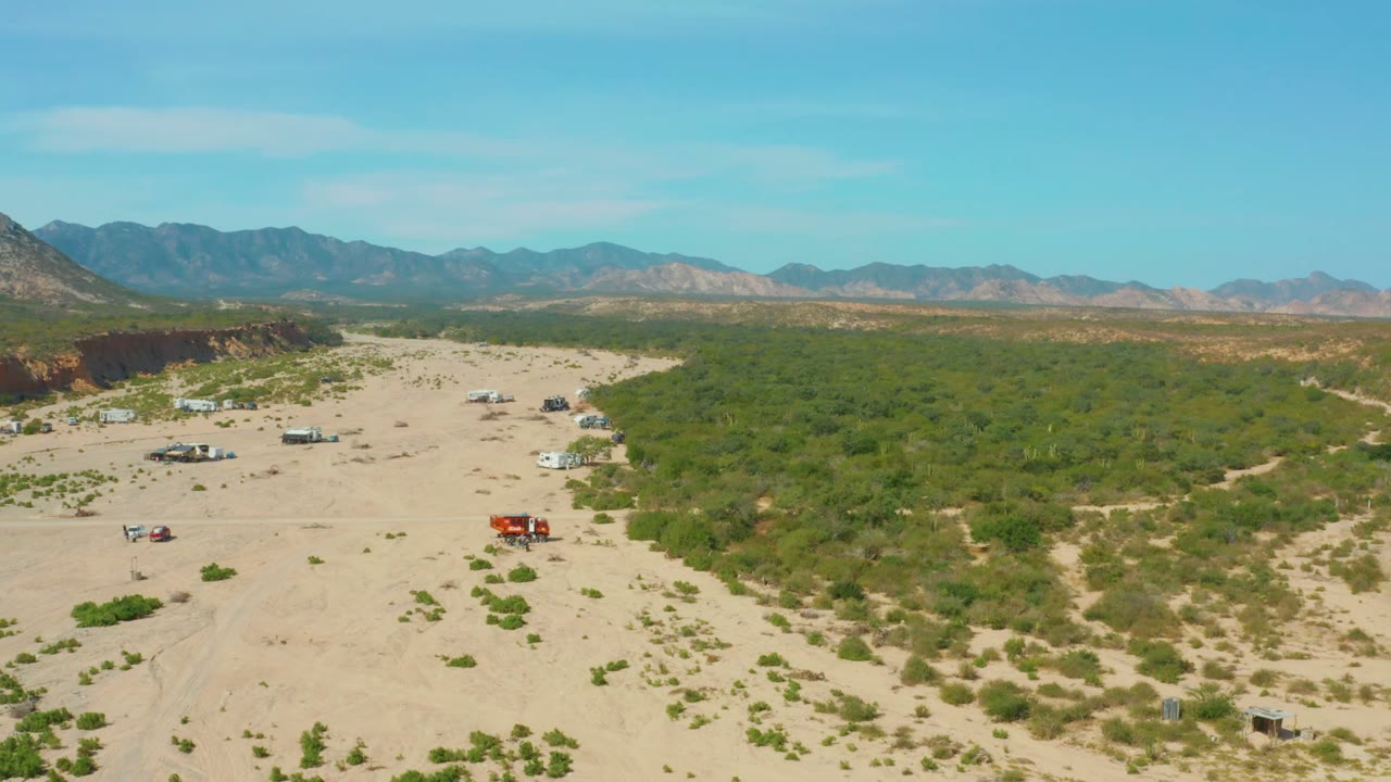 Wide aerial desert scene with vehicles on sand, green shrubs, mountains, and clear sky.