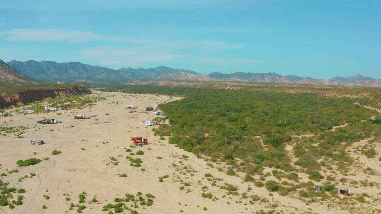 Los Frailes Los Cabos aerial frame showing a red truck on open sand, small parked vehicles nearby, scrubland filling the right side, distant ridges, clear sky, and no visible people.