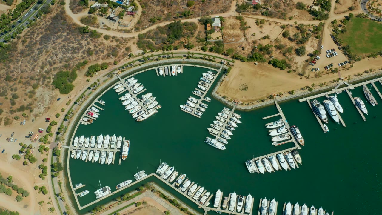 Opening overhead frame of Puerto Los Cabos marina with green water, white docked boats, curved harbor edge, dry tan land, sparse vegetation, and no visible people.