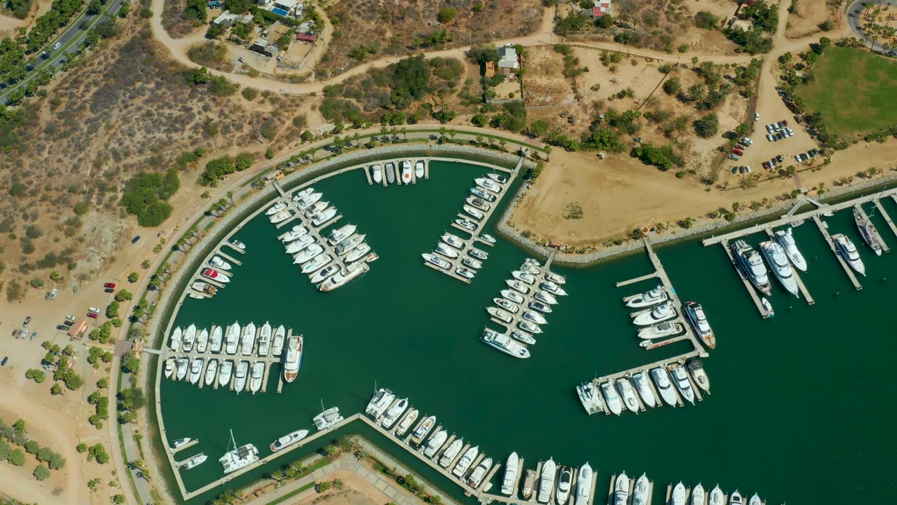 One second into the Puerto Los Cabos aerial, docked yachts cluster around piers in green marina water beside dry shoreline, scattered trees, road edges, and no visible people.