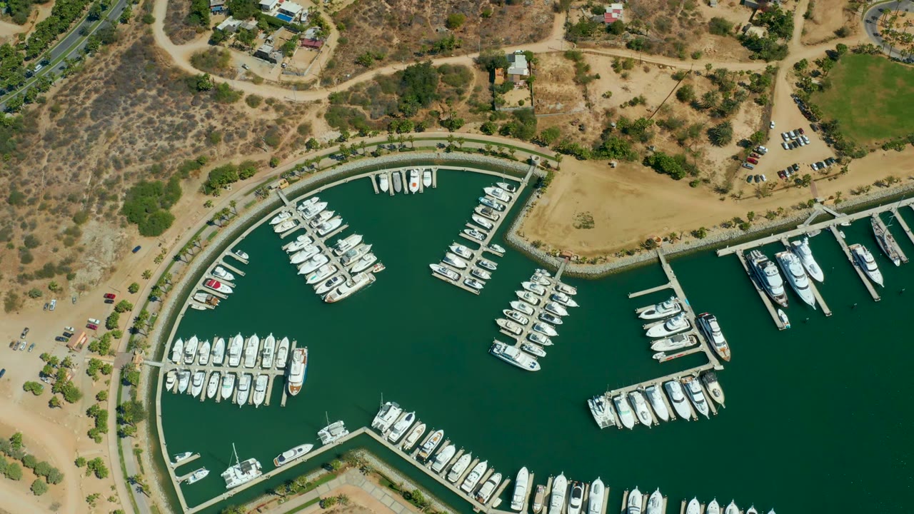 Mid-early frame of Puerto Los Cabos marina showing white boats lined along piers, a curved quay, dry scrubby land above the waterline, parked vehicles, and no visible people.