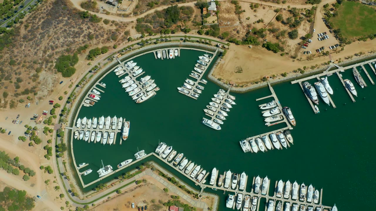 Overhead view of Puerto Los Cabos marina with rows of white boats in green water, curved docks, tan dirt roads, dry hillside terrain, parked vehicles, and no visible people.