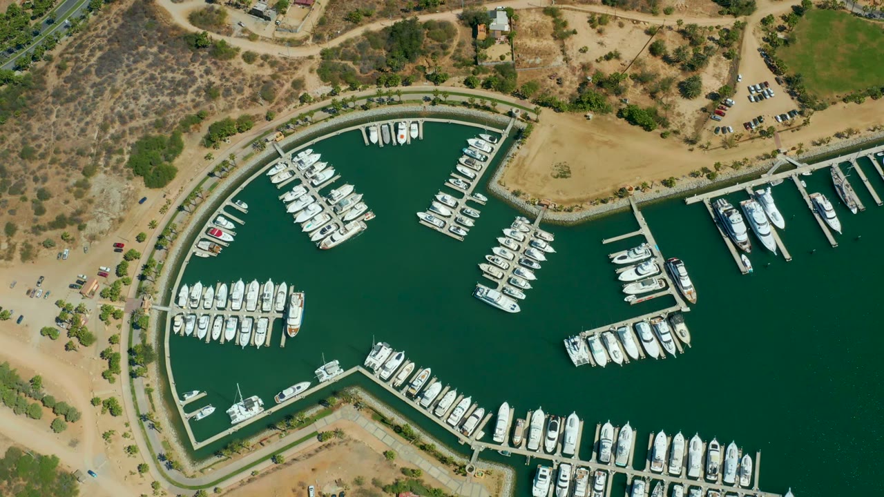Aerial marina view with clustered boats, docks, green water, parked cars, and dry land.