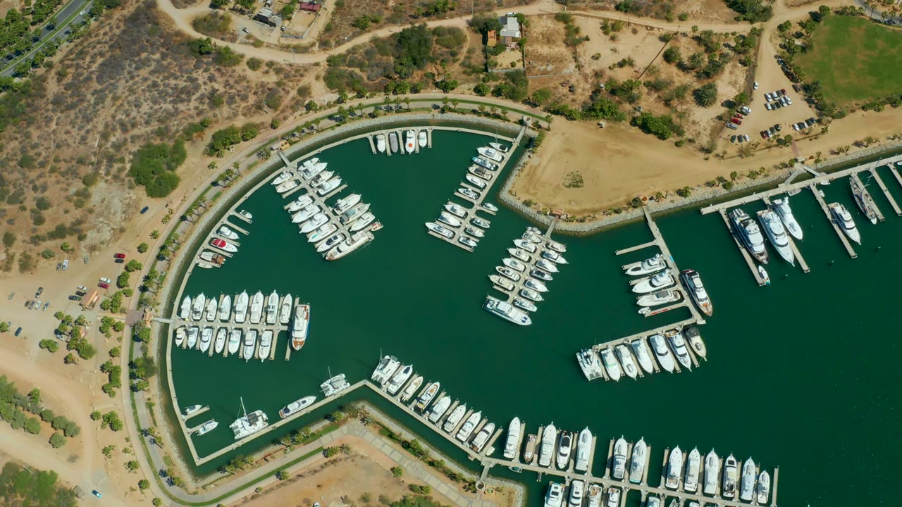 At 0:02, a top-down Puerto Los Cabos marina frame shows white yachts arranged around dock fingers, green water, tan access roads, dry scrubby hills, a green field edge, and no visible people.