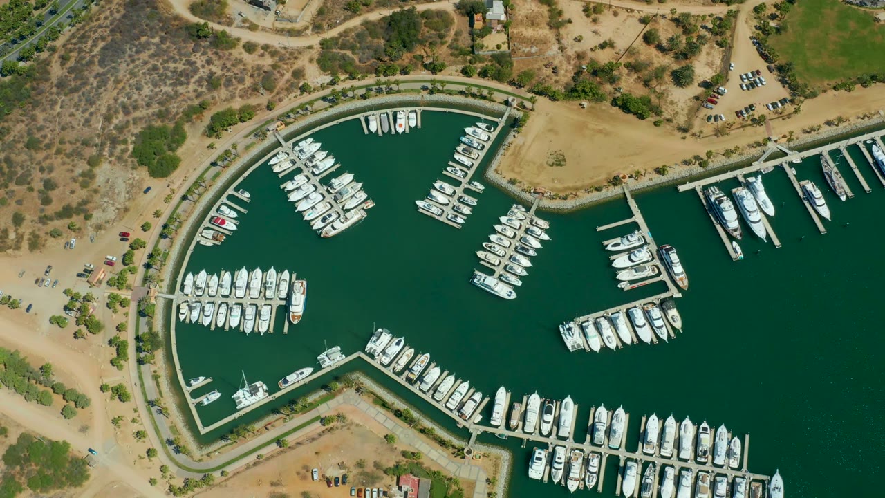 At two seconds, the Puerto Los Cabos marina shows clustered boats on the left and lower slips, open green water on the right, tan land at the top, and no visible people.