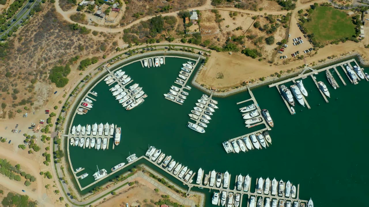 Opening overhead view of Puerto Los Cabos marina, with white boats arranged along docks, green harbor water, sandy shore, dry hills, parked vehicles, and no visible people.