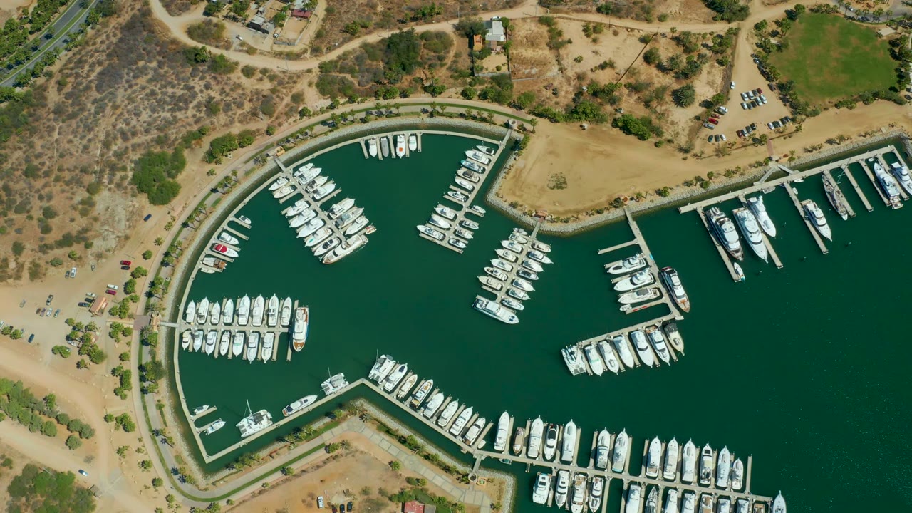 Mid-clip overhead frame of Puerto Los Cabos marina with dense rows of white boats, green water, dry sandy shoreline, hillside brush, parked cars, and no visible people.