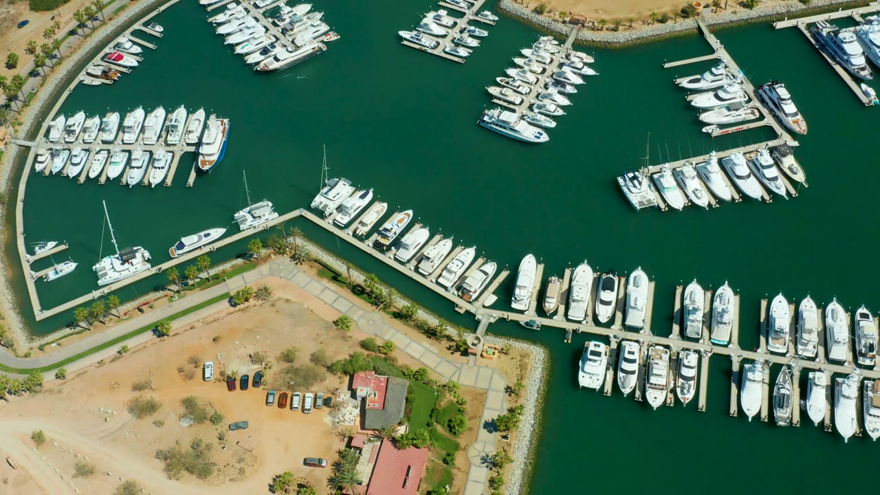 Overhead aerial frame of Puerto Los Cabos marina with rows of white boats along docks, green water, sandy shore, parked vehicles, red-roof buildings, and no visible people.