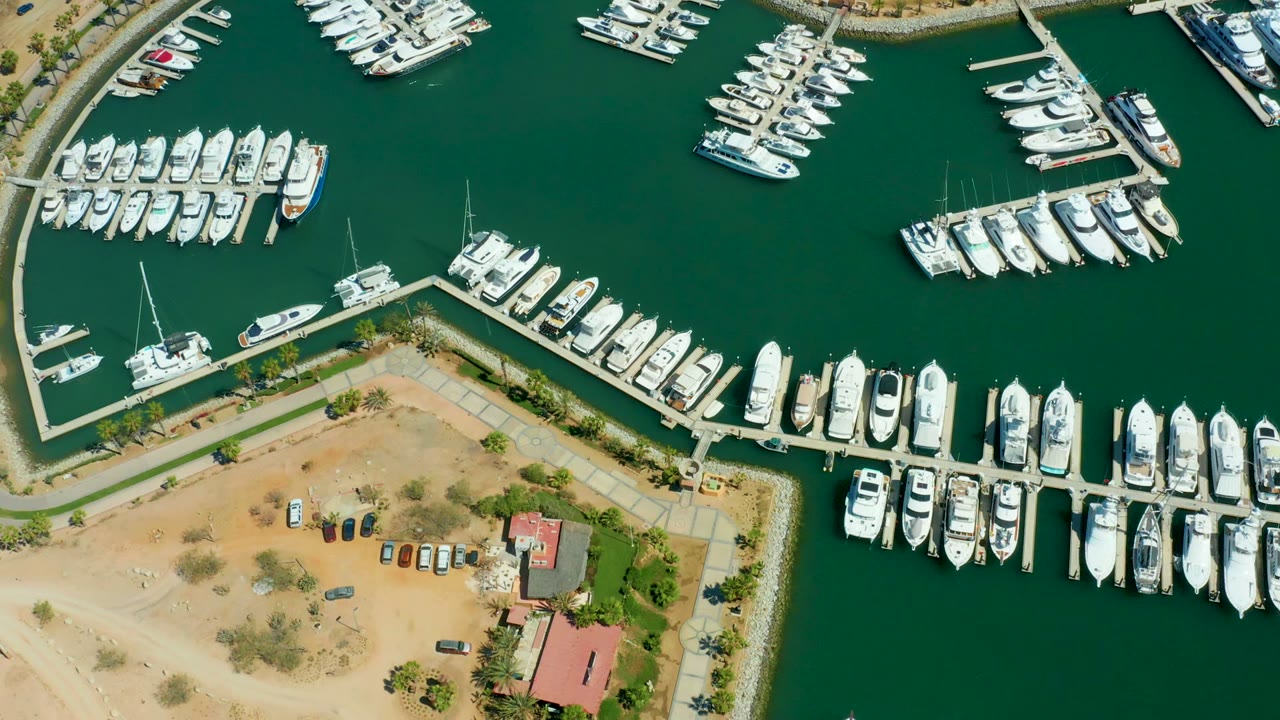 Aerial frame over Puerto Los Cabos marina with boats lining two dock areas, deep green water, a pale sandy shore, small trees, vehicles, and no visible people.