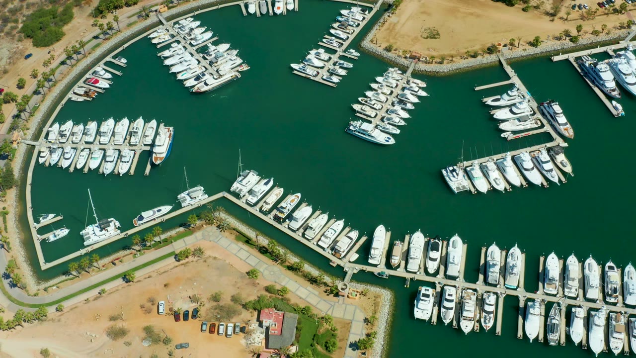 Overhead view of Puerto Los Cabos marina with rows of white yachts and sailboats around angular docks, sandy land at the lower edge, and no visible people.