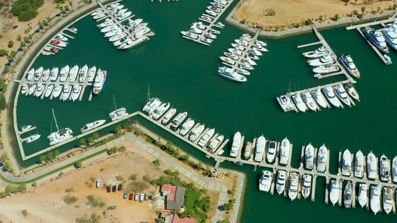 Top-down view of Puerto Los Cabos harbor with white yachts clustered along several docks, deep green water between slips, land at the bottom, and no visible people.