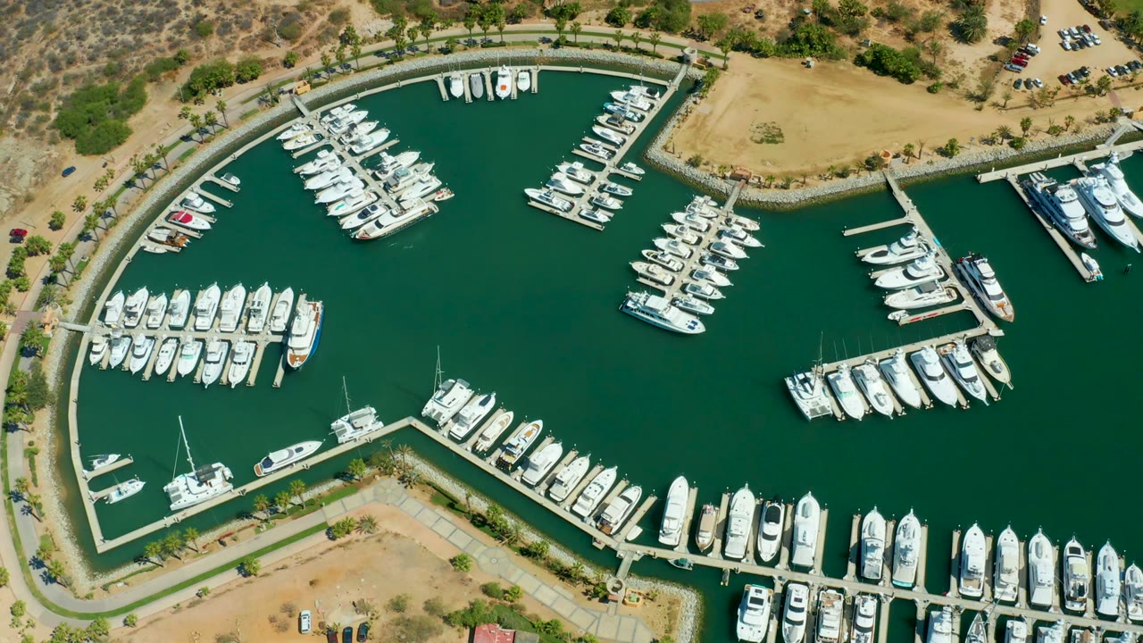 Overhead view of Puerto Los Cabos marina with rows of white boats on docks, green harbor water, dry tan land around the basin, and no visible people.
