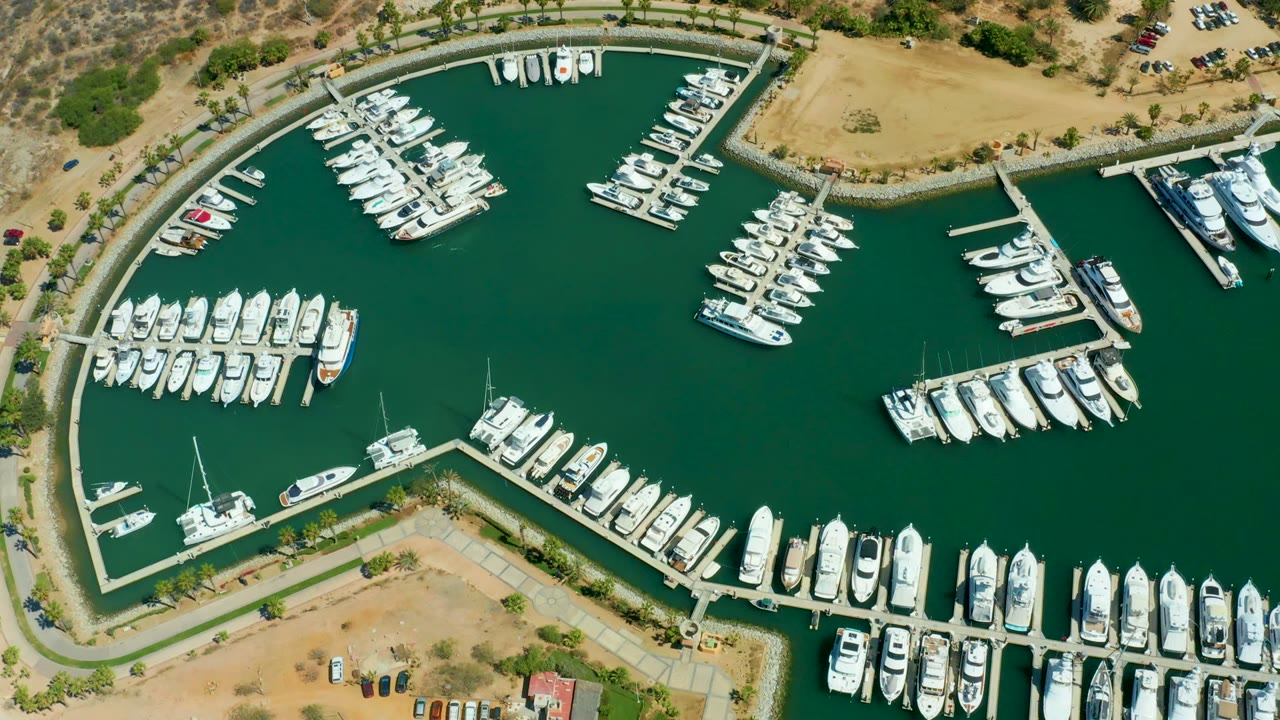 High overhead view in Puerto Los Cabos with curved marina edge, multiple rows of boats, dark green water, dry land, parked vehicles, and no visible people.