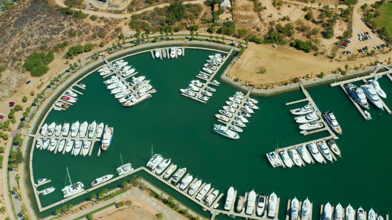 Overhead view of Puerto Los Cabos marina at the start of the clip, with white boats moored along curved docks, green water, tan shoreline, and no visible people.