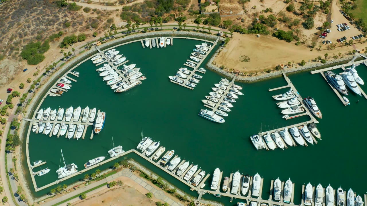 Puerto Los Cabos marina from above at 0:02, showing white yachts along the bottom and right docks, small craft at the upper slips, green water, and no visible people.