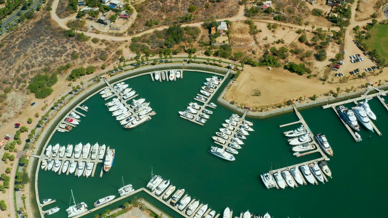 Opening overhead frame of Puerto Los Cabos marina with white boats along curved docks, green water, tan dry ground, roads, parked cars, scattered buildings, and no visible people.