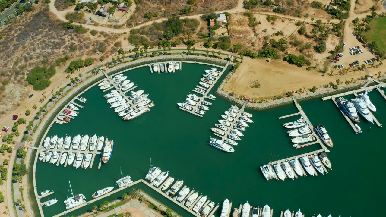At 0:02, the aerial frame shows Puerto Los Cabos boats arranged around multiple docks, a curved seawall, adjacent roads, parked cars, dry scrub terrain, and no visible people.