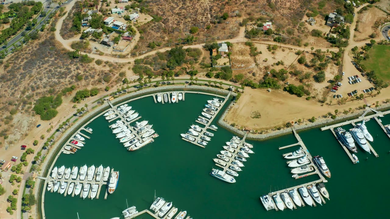 Aerial frame over Puerto Los Cabos marina showing rows of white boats in green water, curved docks, sandy shoreline, dry hills, roads, cars, and no visible people.