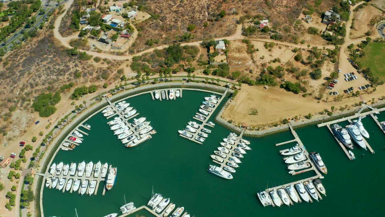 Overhead marina view with docked boats, green water, road, cars, and dry hillside.