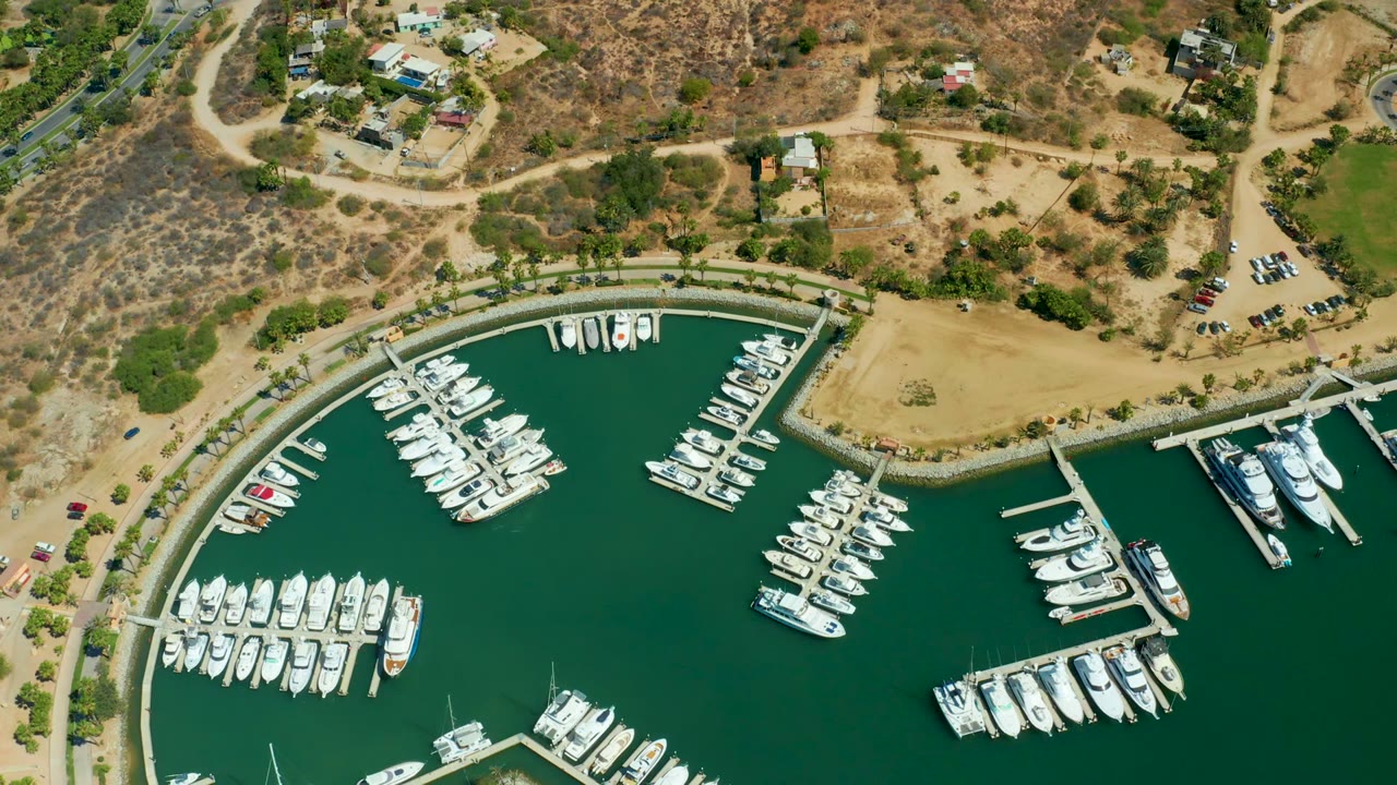 Puerto Los Cabos marina aerial showing yachts arranged along multiple docks, green harbor water, sandy open land, parked cars, dry hills, and no visible people.