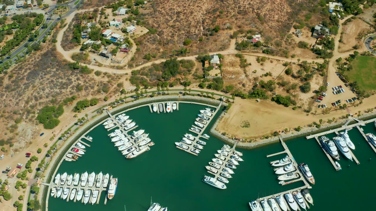 Opening overhead frame of Puerto Los Cabos marina with white boats in green water, sandy shoreline, dry hills, roads, parked cars, hillside homes, and no visible people.
