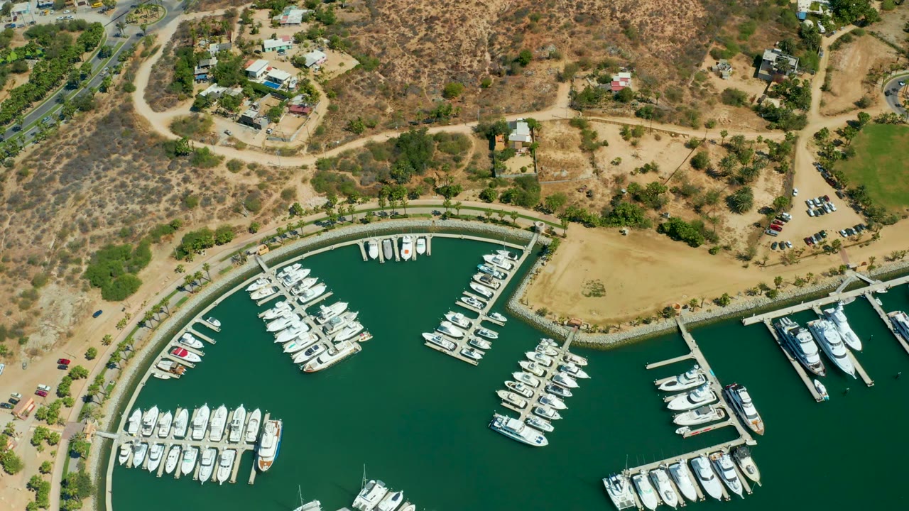 Third frame from above Puerto Los Cabos marina, with rows of docked yachts in dark green water, a shoreline path, parked cars, dry scrub hills, nearby homes, and no visible people.