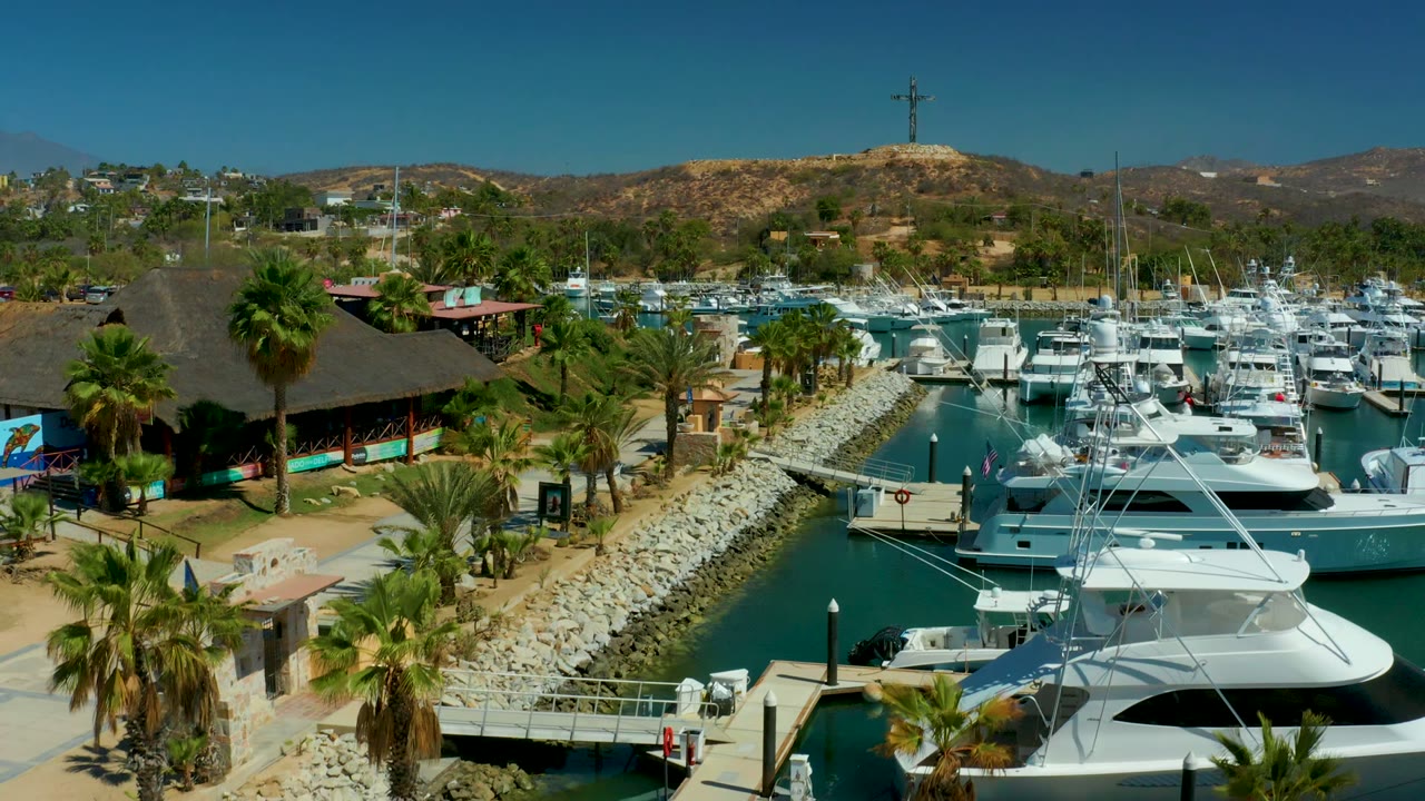 Puerto Los Cabos marina from above with white boats, a palm-lined walkway, a rock-edged water channel, dry hills, and no visible people in this first frame.