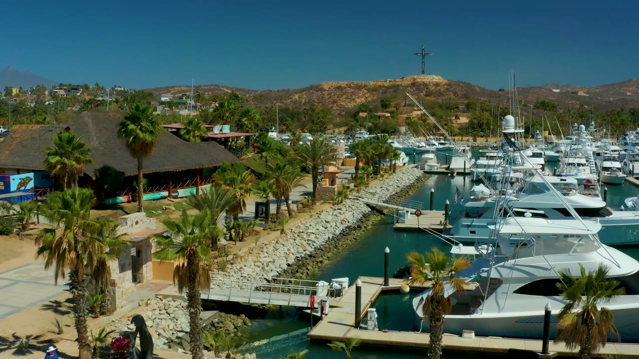 Puerto Los Cabos marina with a large white yacht in the lower right, dock ramps and palms near the water, hills behind, and no visible people in this frame.