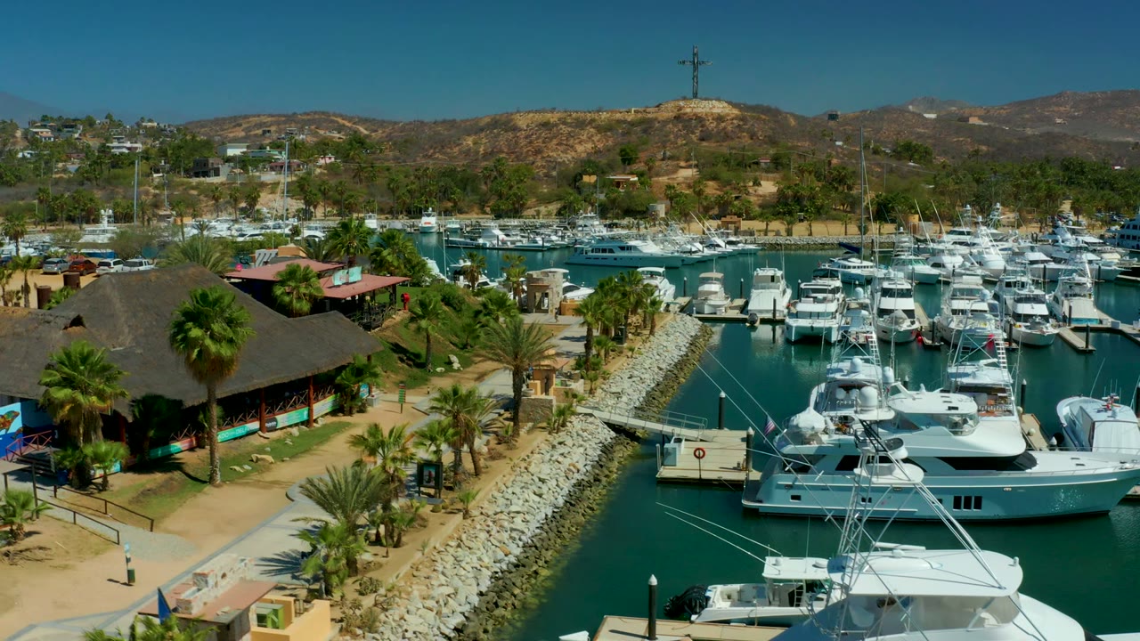 Wide aerial frame of Puerto Los Cabos marina showing white yachts in foreground and midground slips, palms beside a sandy walkway, dry hills, a ridge cross, and no visible people.