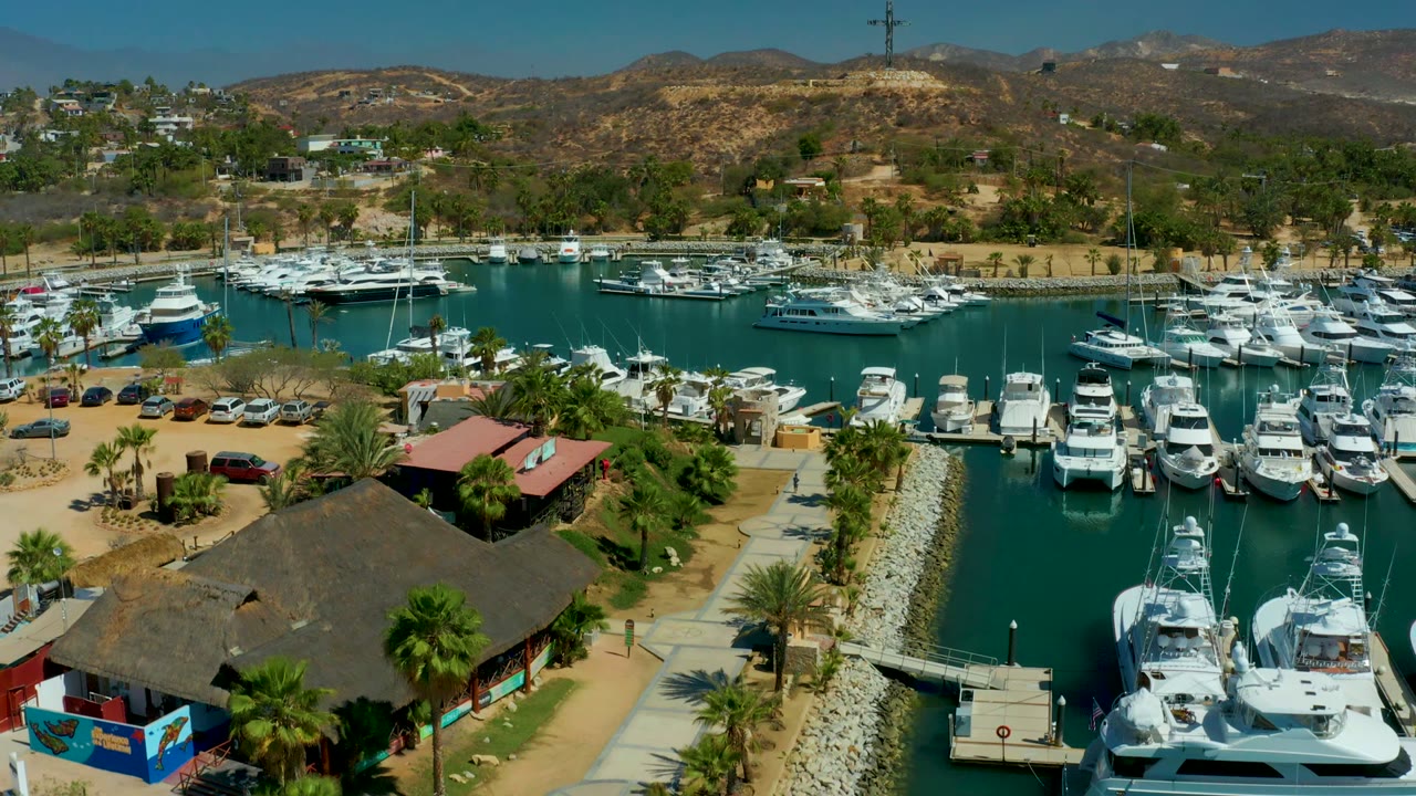 Opening aerial frame over Puerto Los Cabos marina with white yachts, a thatched-roof waterfront building, palms, parked cars, dry hills, and no visible people.