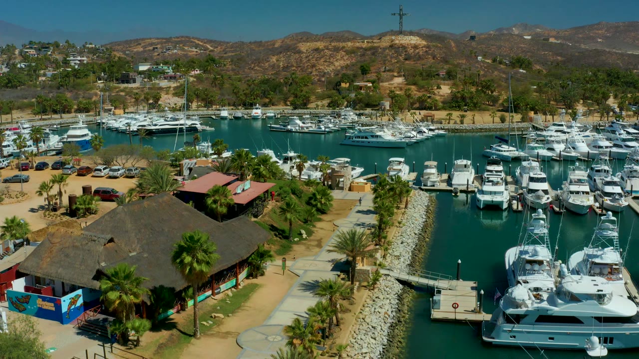 Puerto Los Cabos marina from above with a large dark thatched roof in the foreground, clustered white boats on both sides of the water, arid hills, and no visible people.