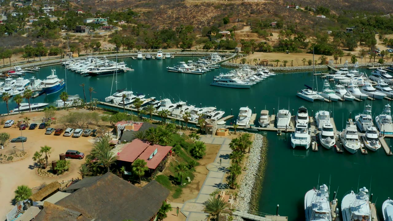 Aerial frame of Puerto Los Cabos marina with docked white boats, green-blue water, a parking lot beside red-roof and thatched-roof buildings, palm trees, dry hills, and no visible people.