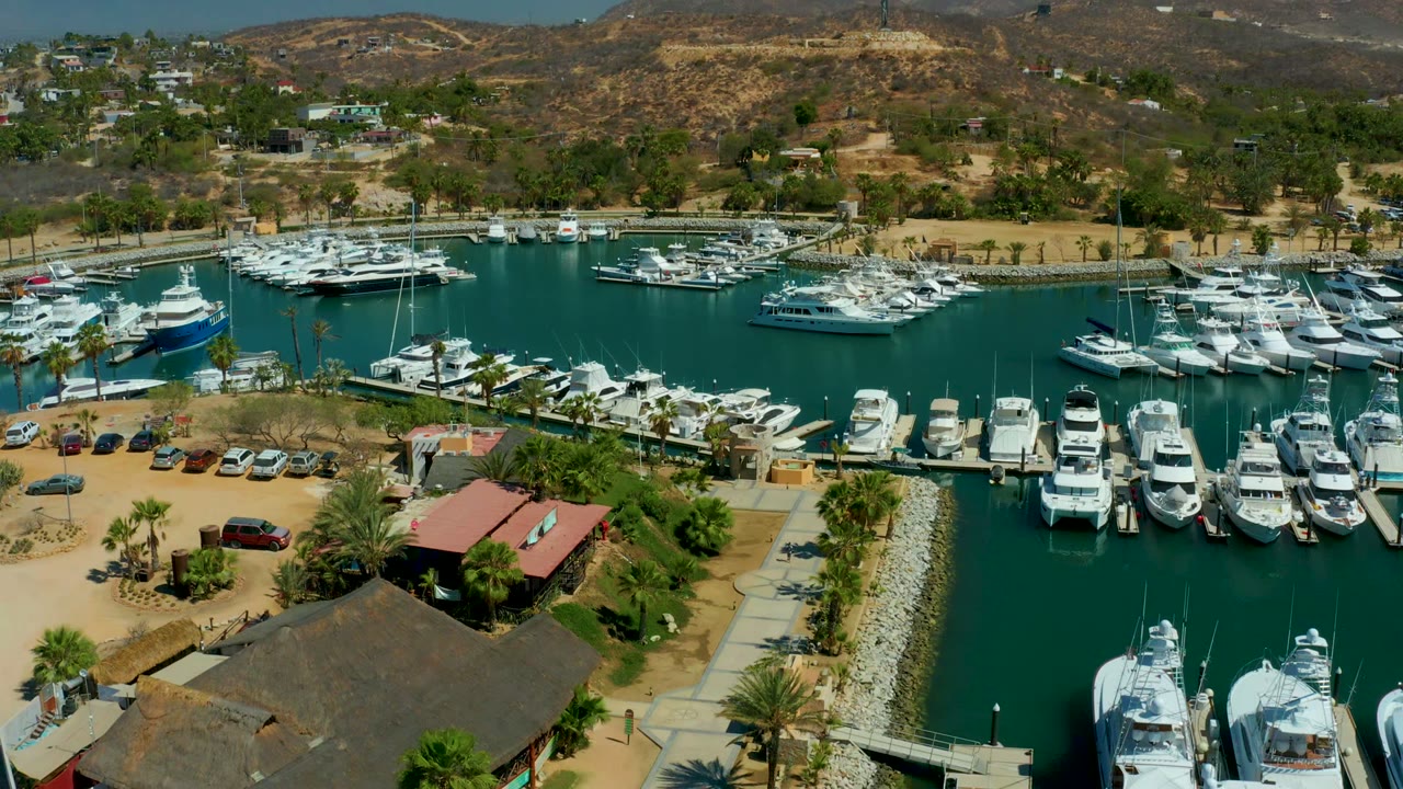 Elevated view of Puerto Los Cabos with marina slips full of white boats, turquoise-green water, waterfront buildings, cars in a sandy parking lot, palms, arid hills, and no visible people.