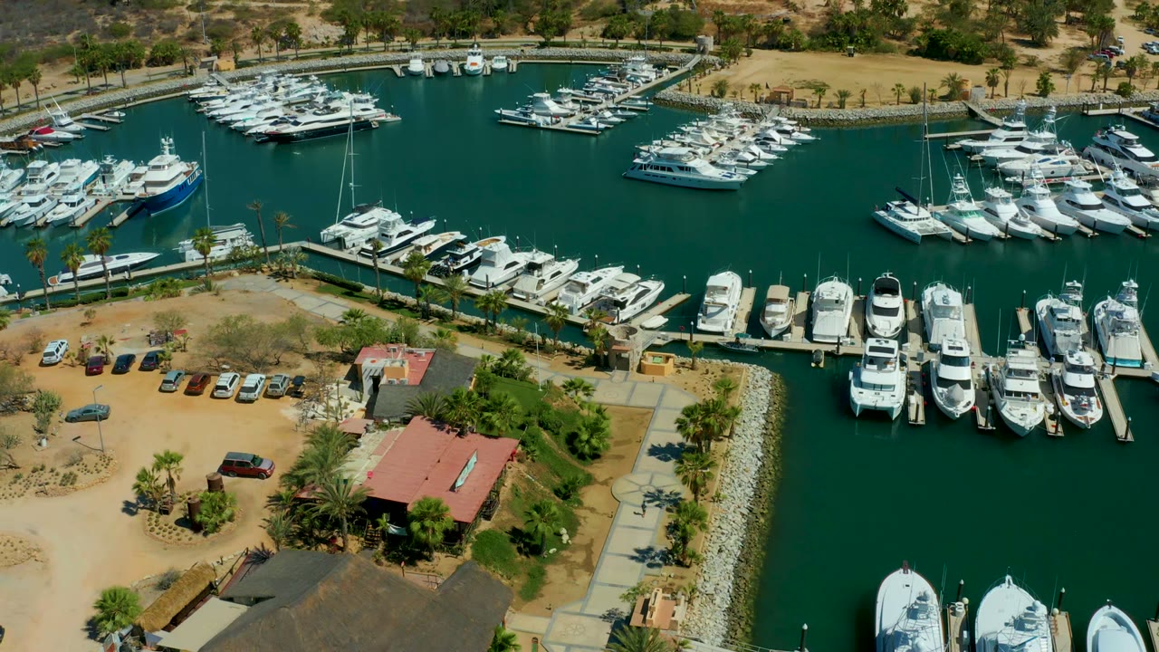 Opening aerial frame over Puerto Los Cabos marina, with white yachts along docks, green water, parked cars, palm trees, dry shoreline terrain, and no visible people.