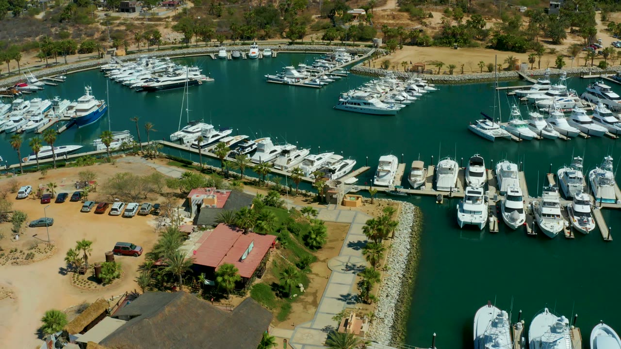 Aerial frame from Puerto Los Cabos marina with white boats packed along piers, green harbor water, a curved shore road area, parked cars, palms, dry hills, and no visible people.