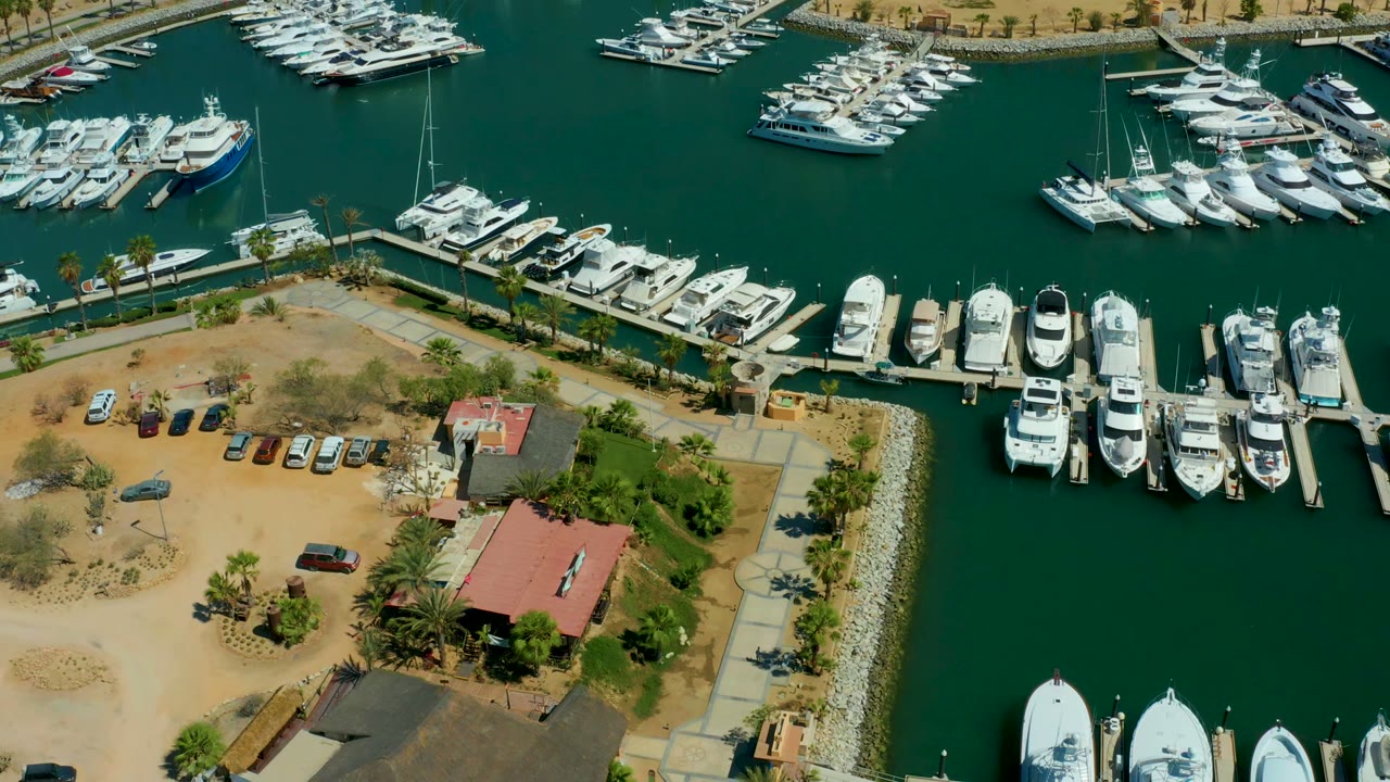 Overhead view of Puerto Los Cabos marina with rows of white yachts, green-blue water, a sandy parking lot, a red-roofed building, palm trees, and no visible people.