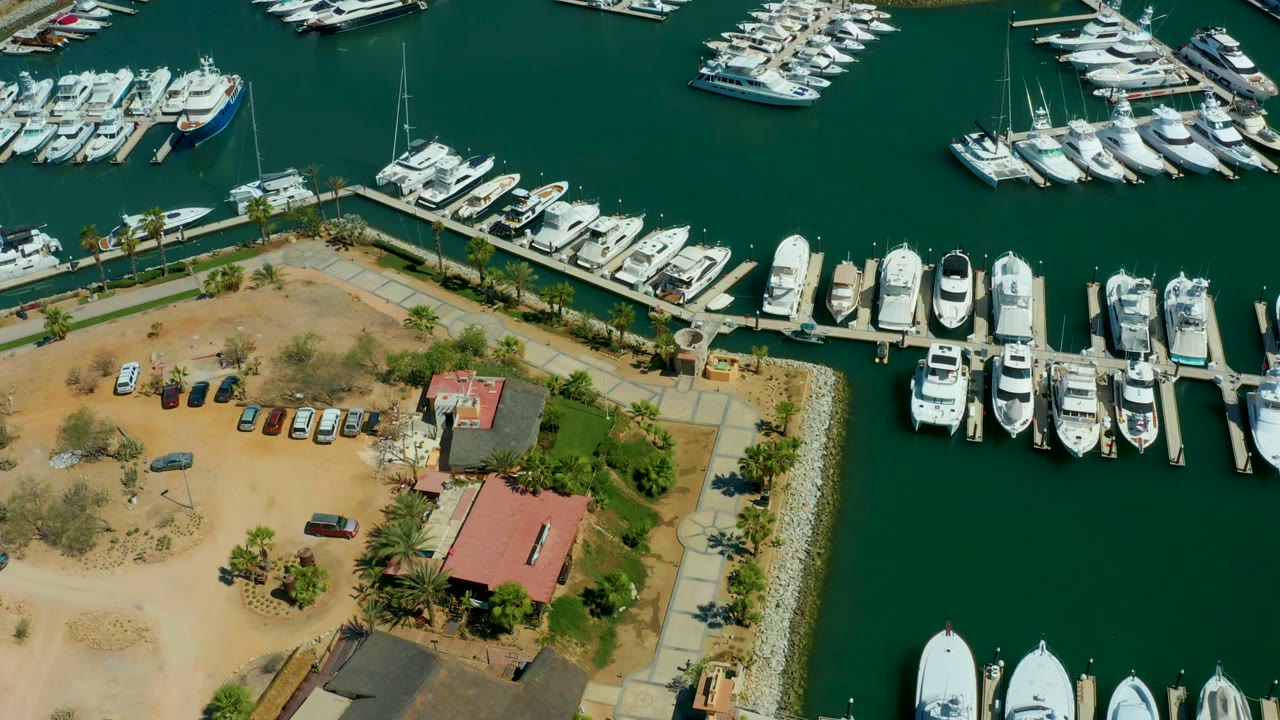 Aerial view of Puerto Los Cabos marina at 0:00, with white yachts along docks, green water, a tan parking lot, red-roofed buildings, palms, and no visible people.