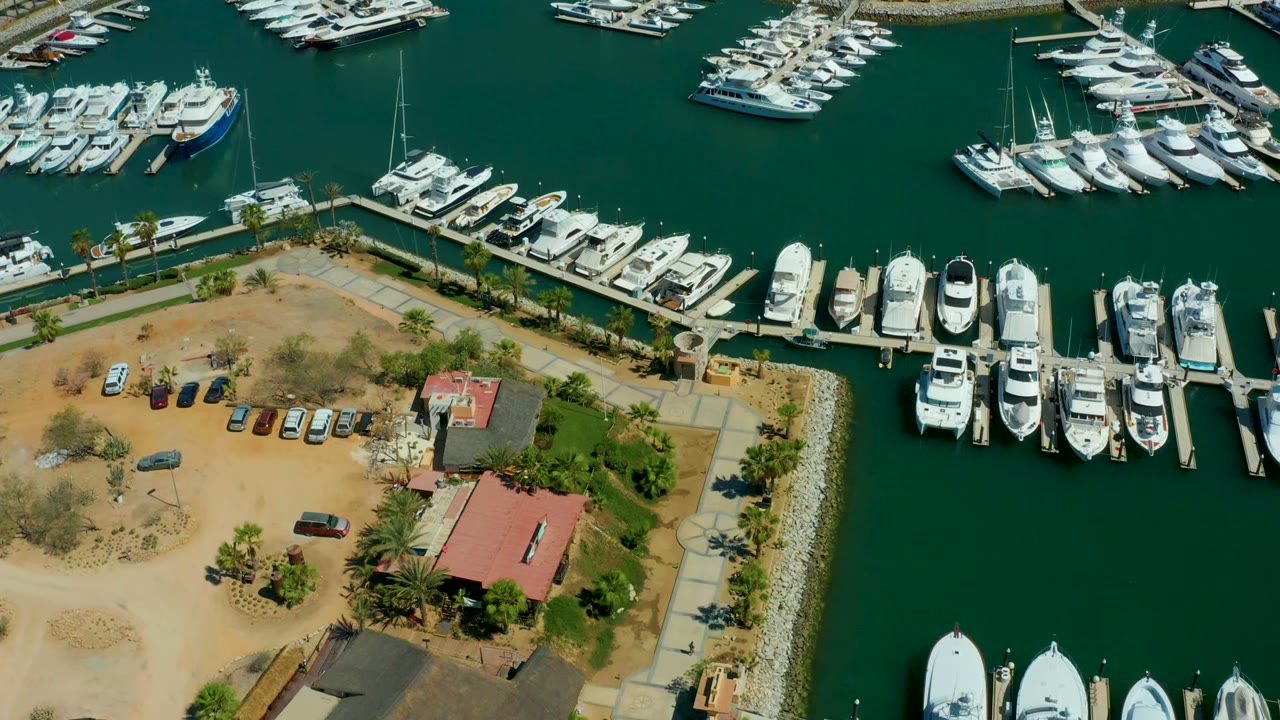Puerto Los Cabos marina at 0:02, with white boats clustered along docks, a waterfront path, parked vehicles on tan ground, red-roofed structures, and no visible people.
