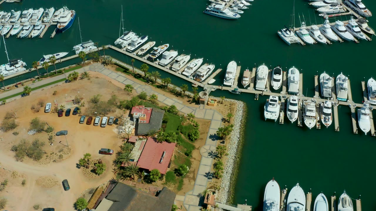 At 0:00, an aerial view over Puerto Los Cabos shows white yachts docked in green marina water beside a dirt parking lot, a red-roof building, palms, and no visible people.