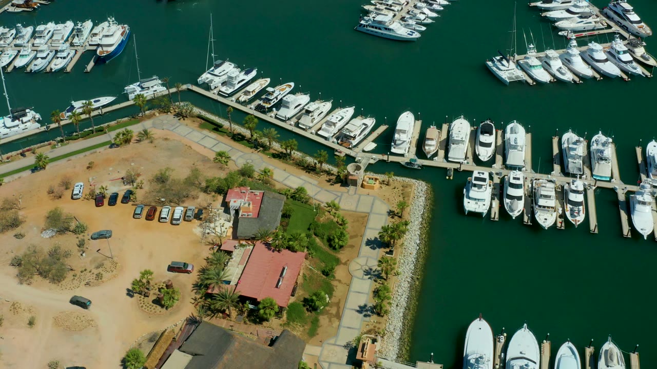 At 0:02, the aerial view over Puerto Los Cabos shows clustered yachts along the docks, green water, the red-roof building, parked cars, and no visible people.