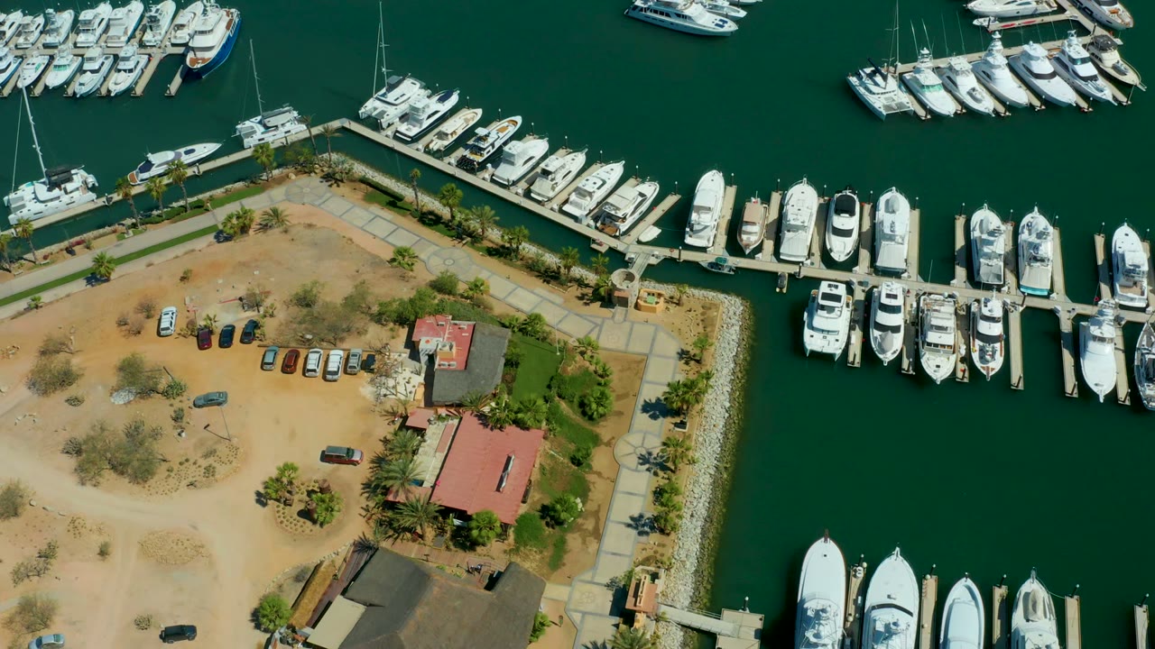 At two seconds, Puerto Los Cabos marina docks form a grid of white boats around dark green water, with sandy land, cars, and no visible people.