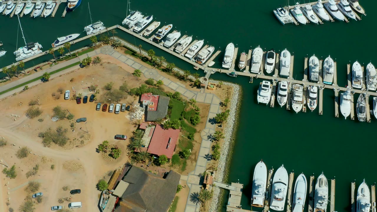 Top-down aerial frame over Puerto Los Cabos marina showing white yachts around docks, a sandy parking lot with cars, red-roof and dark-roof buildings, palm trees, green water, and no visible people.