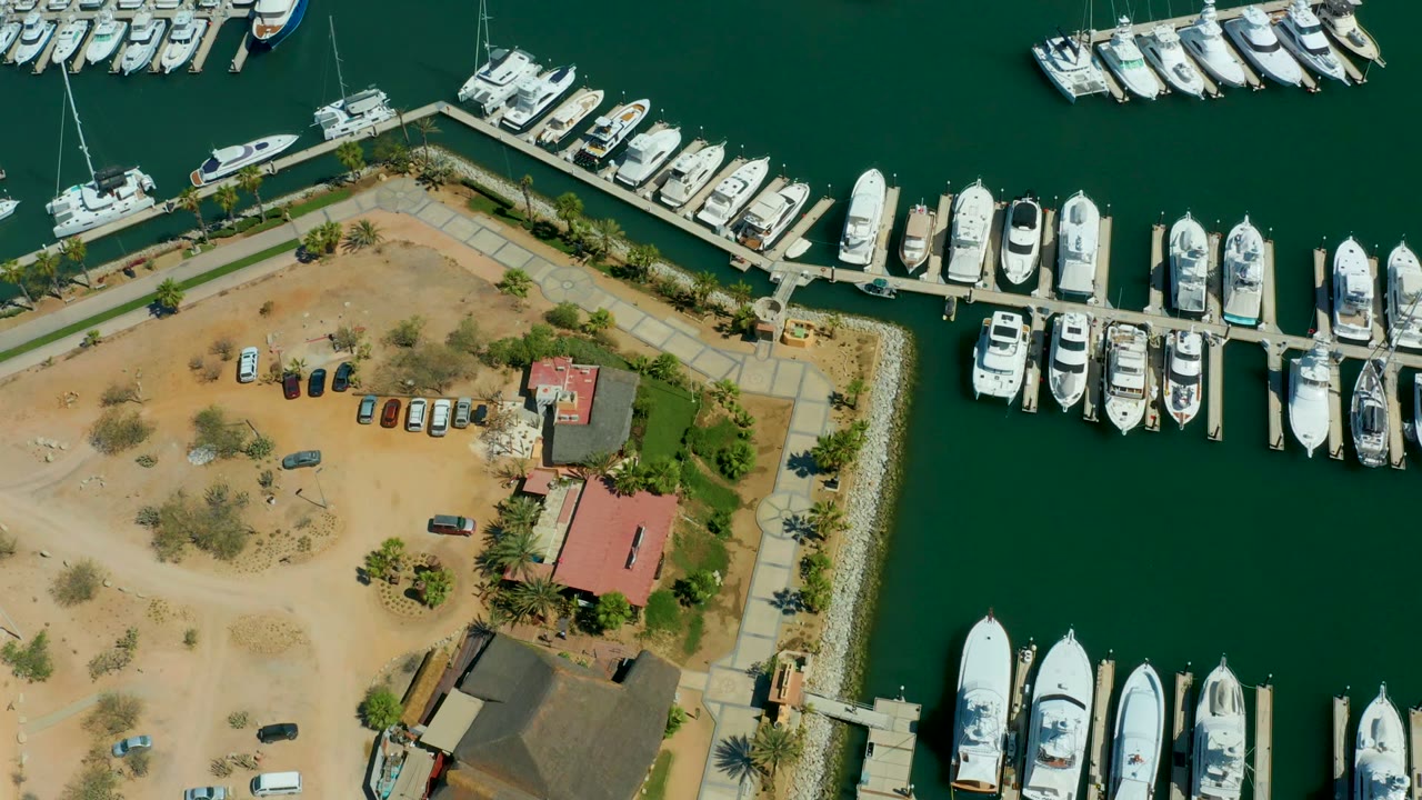 Aerial marina view with docked yachts, scattered cars, palm trees, and waterfront buildings.