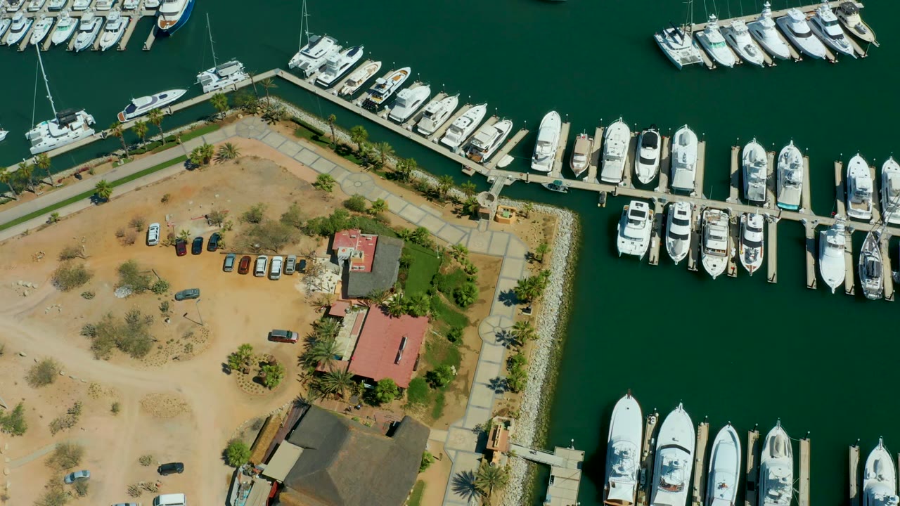 Mid-clip aerial frame at Puerto Los Cabos marina showing a broad band of green water, yacht slips along the docks, cars spread across the sandy parking area, waterfront buildings, and no visible people.