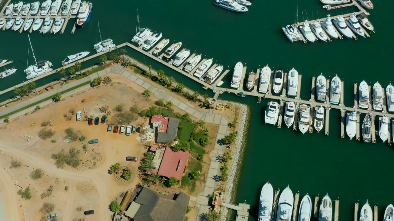 Overhead frame of Puerto Los Cabos marina showing green water, rows of white yachts, sandy parking areas, red-roofed buildings, and no visible people.