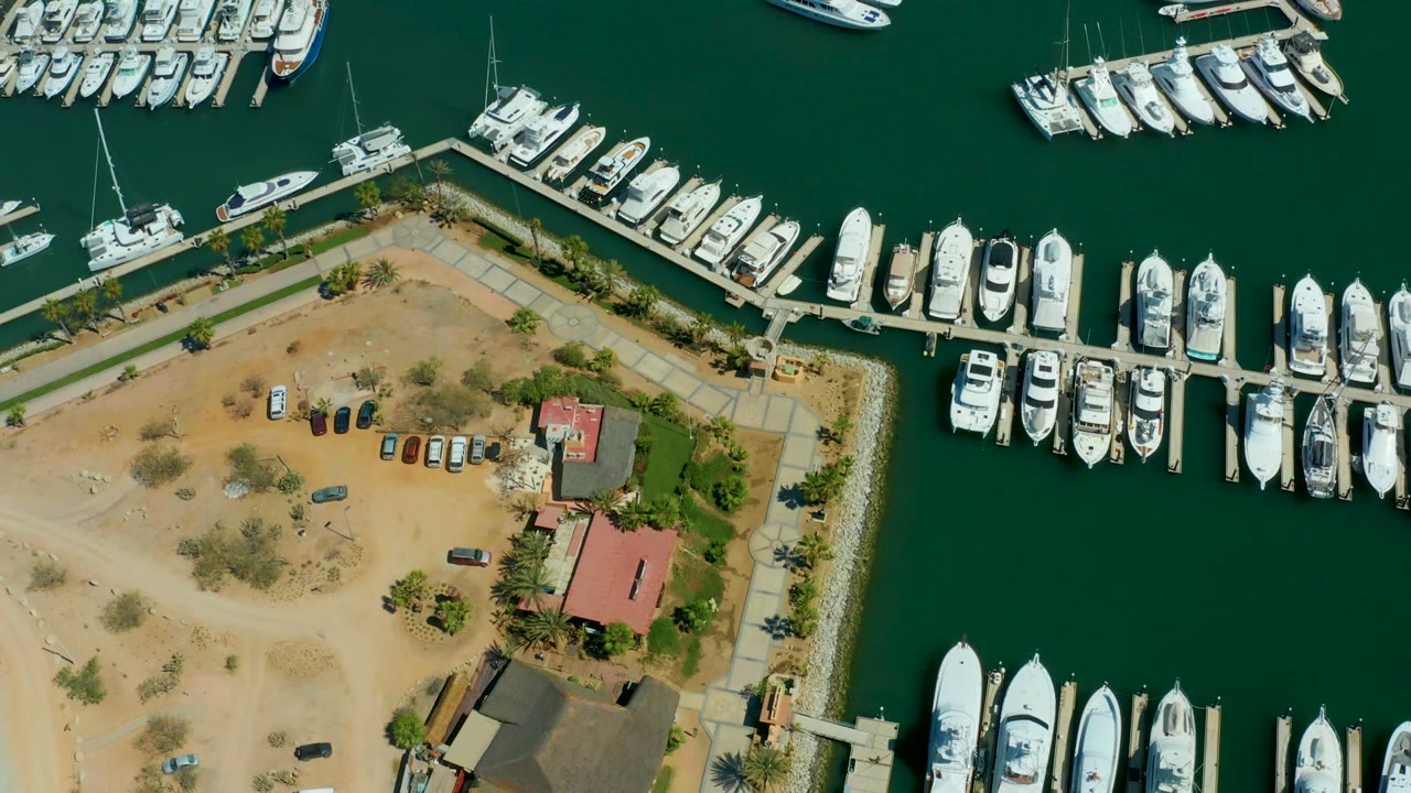 Aerial view of yacht slips beside a sandy lot with cars and red roofs.