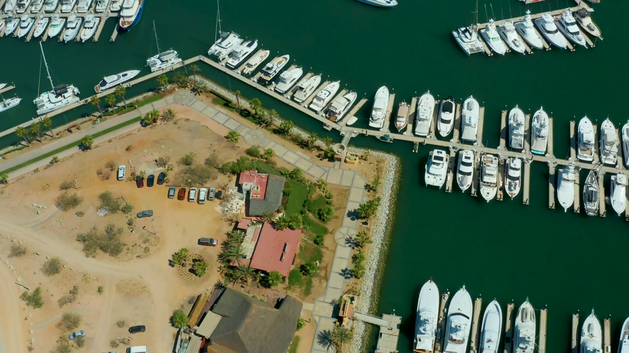 A top-down view over Puerto Los Cabos shows the harbor edge, docked yachts, an open sandy parking area, a red-roofed building, and no visible people.