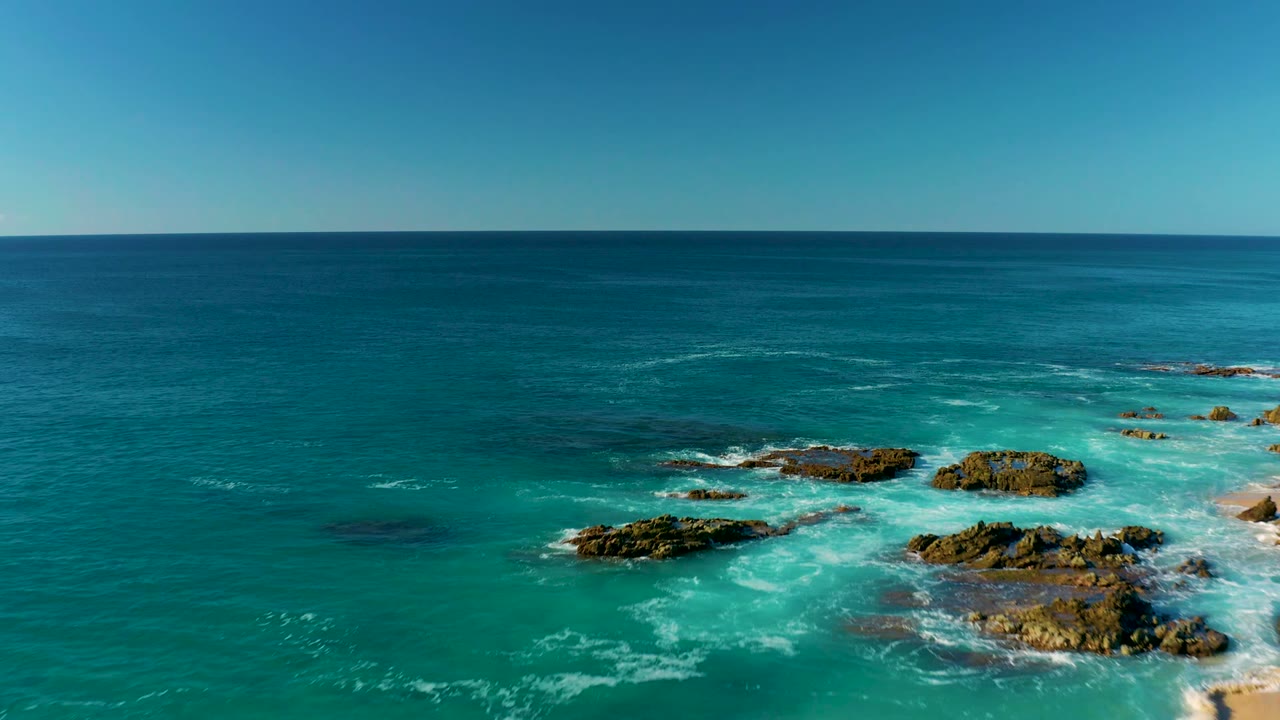 Opening aerial view at La Fortuna, Baja California Sur, with turquoise water and white foam around rocky outcrops in the lower frame and no visible people.